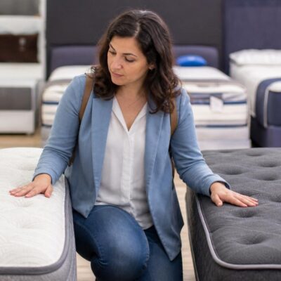 Woman in a blue blazer kneeling between a white and a dark grey tufted mattress, touching both to compare their firmness in a showroom.